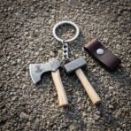 Hand forged viking keyring with miniature damascus axe and hammer charms on ash handles and a brown leather fob lying on wet gravel