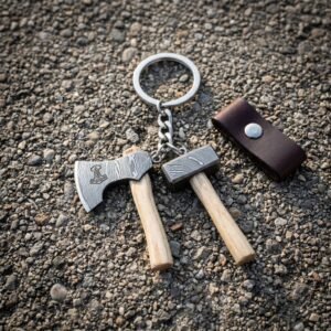 Hand forged viking keyring with miniature damascus axe and hammer charms on ash handles and a brown leather fob lying on wet gravel