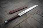 Hand forged Viking langseax with knotwork etching and broken back blade, diagonal view on flagstones next to its fitted scabbard