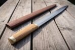 Hand forged Viking seax with olive wood handle and brass ferrule lying on weathered table boards beside its leather sheath