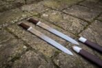 Hand forged Viking seax pair with leather grips crossing on mossy flagstones next to two dark fitted scabbards