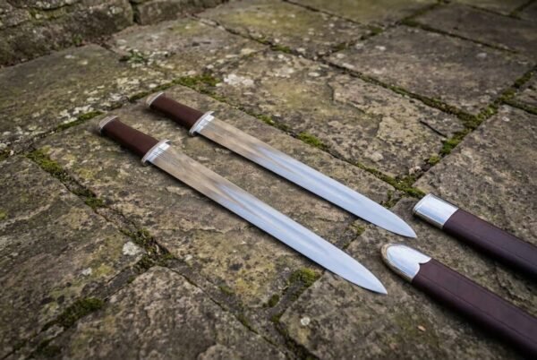 Hand forged Viking seax pair with leather grips crossing on mossy flagstones next to two dark fitted scabbards