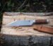 Hand forged viking seax with a broken back blade, etched raven and stacked leather handle lying on a split log beside its hand stitched tan leather sheath