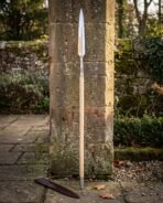 Hand forged viking spear standing against a stone pillar, long steel head with a knotwork cut socket on an ash shaft with a leather blade cover below.