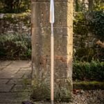 Hand forged viking spear standing against a stone pillar, long steel head with a knotwork cut socket on an ash shaft with a leather blade cover below.