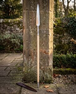 Hand forged viking spear standing against a stone pillar, long steel head with a knotwork cut socket on an ash shaft with a leather blade cover below.