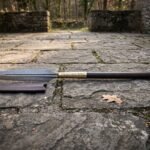 Hand forged Viking spear with a Damascus leaf blade in 1095 and 15N20, rune cut brass socket and dark stained ash shaft lying on flagstones beside its leather sheath