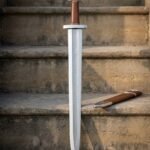 Hand forged viking sword with a pattern welded herringbone fuller and trilobate Valknut pommel, standing on sunlit stone steps beside a brown leather scabbard