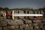 Hand forged viking sword with polished fullered blade, engraved brass knotwork guard, red leather grip and brass ring pommel lying on a dry stone wall