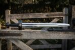 Hand forged viking sword with a Valknut engraved blackened guard and black leather wrapped grip resting on a weathered wooden farm gate beside its dark scabbard
