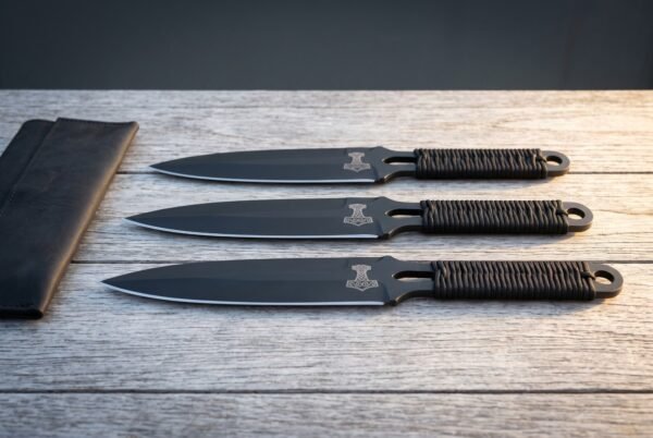 Hand forged Viking throwing knife set of three with black coated blades, etched Thor hammer marks and cord wrapped handles, laid on grey timber beside a leather roll.