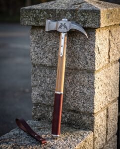 Hand forged war hammer with a Damascus steel head, etched ravens and a curved fluke, ash haft with steel langets and an oxblood leather grip, against a granite pillar.