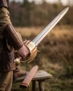 Hand gripping a Damascus short sword by its brass Vegvisir basket hilt with a tan leather scabbard behind