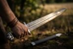 Hand gripping a Damascus Viking sword with lobed steel pommel and etched knotwork fuller over its scabbard
