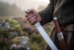 Hand gripping a single edged steel seax with knotwork engraved bolster beside a matching sheath on a leather belt
