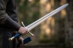 Hand gripping a steel sword with blue leather grip and curved guard, holding the blue scabbard in the other hand