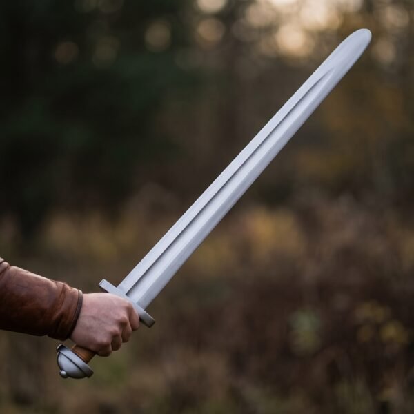Hand holding a blunt steel trainer sword with tan leather grip and rounded tip against a blurred autumn wood