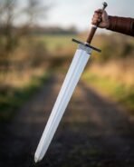 Hand holding a broad steel arming sword with brown wrapped grip, black crossguard and knotwork band on a country lane
