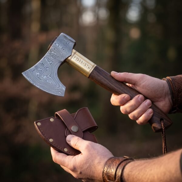 Hand holding a Damascus bearded axe with brass rune collar and wood haft, other hand holding the leather head sheath