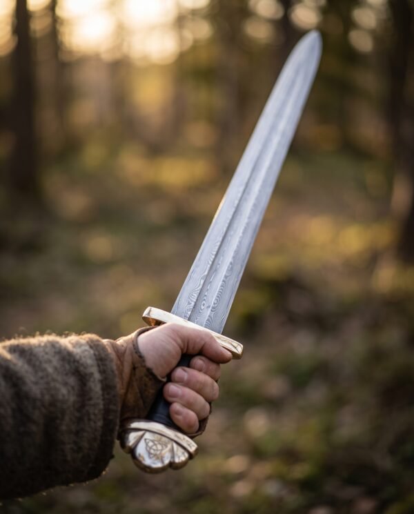 Hand holding a Damascus steel Viking sword with brass guard and triquetra pommel up in a sunlit forest