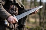 Hand holding a fullered Norse sword with engraved knotwork guard and lobed pommel against a blurred forest