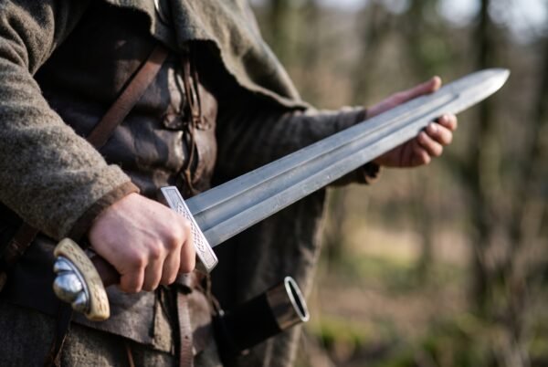 Hand holding a fullered Norse sword with engraved knotwork guard and lobed pommel against a blurred forest