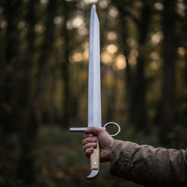 Hand holding a langes messer trainer upright in a dark pine forest, ring nagel beside the hand and beak shaped cap below