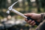 Hand holding a polished steel claw hammer with rune engraved hickory handle and black grip against a blurred forest