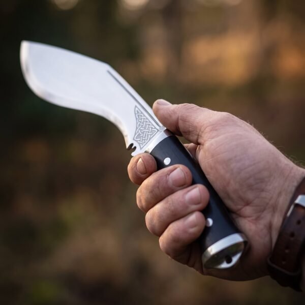 Hand holding a polished steel kukri with knotwork etch and black micarta handle against autumn trees