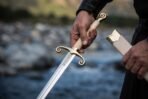 Hand holding a steel sabre by its ribbed pale grip, showing the file worked forte and scrolled brass guard over a stream