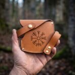 Hand holding a stitched tan leather axe sheath stamped with a Vegvisir and fitted with brass snap fasteners in a forest