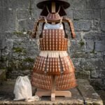 Hand laced leather lamellar samurai armor on a wooden stand against a stone wall, showing wide brim helmet, shoulder guards, rune engraved chest plate and thigh guards.