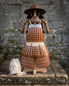 Hand laced leather lamellar samurai armor on a wooden stand against a stone wall, showing wide brim helmet, shoulder guards, rune engraved chest plate and thigh guards.