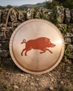Hand made bronze faced hoplon shield, a Greek aspis about 30 inches across with a riveted rim, painted running boar device and leather carry strap against a dry stone wall