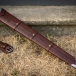 Hand made brown leather sword scabbard with a stamped Valknut, steel throat and chape and a separate belt frog lying on dry grass beside a stone step