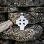 Hand made celtic cross pendant with a pierced ring, brushed steel tone face and stamped Elder Futhark runes hanging from a black leather cord on a lichen covered stone wall