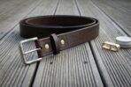 Hand made dark brown full grain leather rune belt with brass rivet, rolled into a coil on grey decking, buckle facing forward