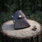 Hand made dark brown leather axe sheath with a tooled Mjolnir panel and a steel snap strap, standing on a cut tree stump beside spare studs and a buckle.