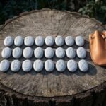Hand made Elder Futhark rune set of twenty four engraved pale stones laid in three rows on a tree stump beside a tan leather drawstring pouch.