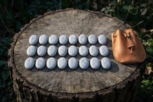 Hand made Elder Futhark rune set of twenty four engraved pale stones laid in three rows on a tree stump beside a tan leather drawstring pouch.