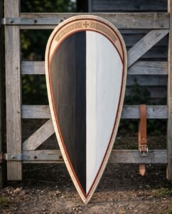 Hand made kite shield with a black and white painted face, rust red trim, riveted rawhide rim and an engraved knotwork panel, leaning against a weathered timber gate.