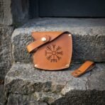 Hand made leather axe sheath in russet vegetable tanned hide with a stamped Vegvisir, brass snap strap and rivets, resting on stone steps beside a leather belt loop