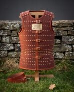 Hand made leather cuirass in russet cowhide with rows of brass domed rivets, five buckle straps and an engraved rune plaque, displayed on a wooden stand by a stone wall
