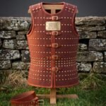 Hand made leather cuirass in russet cowhide with rows of brass domed rivets, five buckle straps and an engraved rune plaque, displayed on a wooden stand by a stone wall