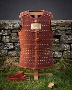 Hand made leather cuirass in russet cowhide with rows of brass domed rivets, five buckle straps and an engraved rune plaque, displayed on a wooden stand by a stone wall