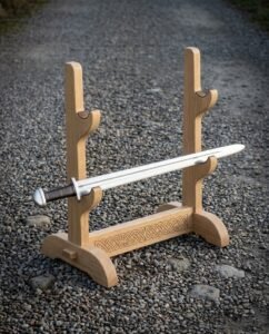 Hand made oak sword display stand with three tiers of cradles and a knotwork engraved crossbar, holding a single polished sword, standing on a gravel path.
