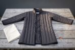 Hand made padded gambeson in charcoal cotton canvas with vertical channel quilting, standing collar and brass grommet cord lacing laid flat on a weathered grey timber table