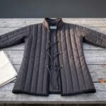 Hand made padded gambeson in charcoal cotton canvas with vertical channel quilting, standing collar and brass grommet cord lacing laid flat on a weathered grey timber table