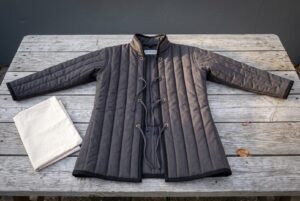 Hand made padded gambeson in charcoal cotton canvas with vertical channel quilting, standing collar and brass grommet cord lacing laid flat on a weathered grey timber table