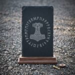 Hand made runic slate plaque engraved with a knotwork Mjolnir inside a full Elder Futhark rune ring, standing in a solid walnut base on grey pebbles.